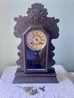 Full view of the antique Ansonia mantle clock featuring detailed wood carvings, visible clock face with Roman numerals, along with the pendulum and winding key displayed in front.