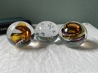 Three art glass paperweights on white cloth showing amber swirl design, bubble-filled clear design, and another amber swirl paperweight.