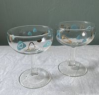 Two atomic boomerang patterned coupe cocktail glasses with turquoise and gold designs on clear stems and bases, shown side by side on white cloth.
