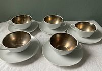 Five pairs of white finely textured linen china tea cups with gold metallic interiors and matching saucers arranged on a table.