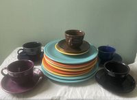 Set of ceramic dishes stacked and arranged including 8 plates, 6 saucers, and 5 cups in various colors such as turquoise, orange, yellow, brown, purple, black, and blue.