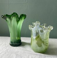 Photo of two green glass vases on a white surface against a green wall; one light green and the other darker green and white swirl, both with ruffled tops.