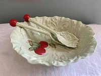 Top front angled view of pale green lettuce leaf shaped bowl with paired lettuce leaf shaped servers inside, showing tomato decoration on bowl and red ends on servers.