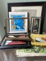Photo showing stacked various framed prints and posters including a yellow ornate frame, a woodblock print of a young girl in a wooden frame, and a plastic picture frame with multiple prints.
