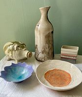 Five pottery items displayed together on white cloth: tall vase, piggy bank, blue bowl, square container, and plate