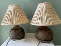 Two pottery lamps with pleated shades, showing round bases and brown earth-tone color palette with incised and raised landscape and building designs