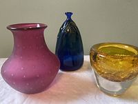 Front view of three signed art glass pieces: pink frosted vase, blue bud vase, and amber crackled vase.