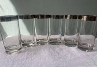 Six clear glass highball glasses with silver rims arranged in a row showing front view and condition of silver tops.