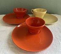 Three snack plates with matching cups, two burnt orange sets and one pale yellow, presented on a white cloth background.
