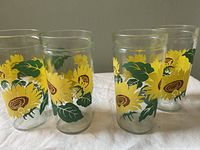 Four clear glass jam jars decorated with bright yellow sunflowers, green leaves, and brown centers, arranged in a row showing front and partial side views.