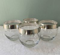 Set of 4 round glass roly poly style glasses with silver metal bands around rims viewed from side and front, arranged in a square.