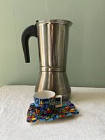 Photo showing full view of the stainless steel espresso maker alongside a Gaudi style small colorful espresso cup placed on its matching square saucer.