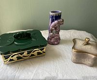 Green trinket box with brass-colored frame, ceramic with lid showing chip damage, next to purple pottery vase and pink powder box.
