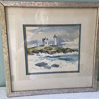 Framed watercolor painting showing overall piece with lighthouse and buildings on rocky shore.