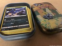 Photo showing a stack of over 40 Pokémon trading cards inside a metal case decorated with a Pokémon design on the lid.