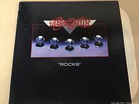 Photo showing the front cover of the Aerosmith vinyl LP titled 'Rocks'. The cover is black with metallic hex bolts and the Aerosmith logo.