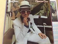 Album cover showing Elton John in white suit sitting by a grand piano, indicating the LP's Greatest Hits album