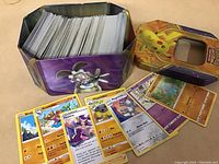 Photo showing a metal tin with a Mega Mawile image containing over 450 Pokémon cards, with the lid featuring Pikachu placed next to it. Several cards laid out in front include Psychic-type and Trainer Fantina cards.