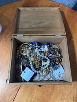 Wooden box open filled with assorted jewelry consisting of pearl necklaces, gold-tone chains, blue bead necklaces, floral decorative pieces, and mixed bracelets.