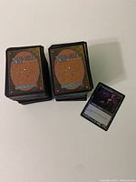Two stacks of Magic: The Gathering cards with backs facing up and one card face up showing artwork of a dark-themed character.