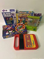 Lot arrangement showing Speak & Spell, two Looney Tunes puzzles, The Brain Train game, Storm game, and handheld water ring toss toy.