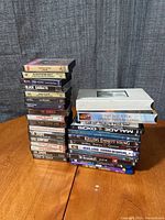 Stack of audio cassette tapes and video game with VHS and DVD cases on a wooden table against a gray curtain backdrop.