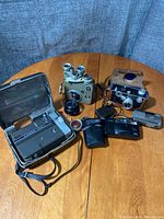 Wide shot showing all vintage cameras and accessories arranged on a wooden table, including the Nikkorex Zoom 8 in open carrying case, assorted cameras, lenses, and cases.