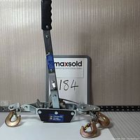 Front view of the full cable puller showing handle with rubber grip, metal body, measuring scale and two hooks with yellow finish.