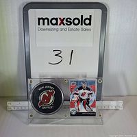 Overall shot of the acrylic display case containing the New Jersey Devils puck and Martin Brodeur card with a MaxSold board in background.