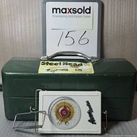 Green TackleMaster solid steel tackle box with metal handle and dent on top. White color-coded Anon Leader weigh scale with circular dial placed in front of the box.