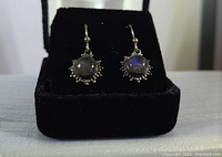 Close-up image of pair of sterling silver earrings each with a round labradorite gemstone center and decorative silver detailing around edges with hook ear wires, displayed on black velvet cushion.