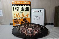 Pair of two used metal signs, one rectangular with orange and black Harley-Davidson text and motorcycle image, and one oval black sign with skull and wing Harley-Davidson logo.