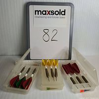 Three sets of older feathered darts in clear plastic cases, each with three darts. One case labeled 15 grams, other two labeled 23 grams. Darts have metal barrels and steel tips with colored feather flights.