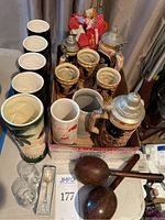 Ceramic beer steins packed in a box, including steins with metal lids and boot-shaped stein. In front are two clear etched shot glasses and a boxed set of souvenir spoons. A pair of wooden maracas is placed beside them and some small flags are seen behind in the box.