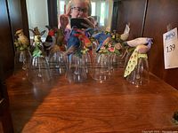 Front view of nine clear glass bells topped with various painted porcelain birds arranged in a row on a wooden surface, showing the intricate details on the birds.
