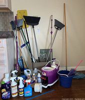 Wide view of assorted brooms, mops, buckets, and various cleaning product bottles grouped on floor against wall.