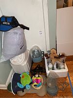 Photo of dog accessories including blue service dog harness attached to white padded bag, plastic water dispenser, food dishes, plush toys, and black feeding dish.