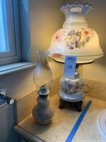 Photo showing two lamps on marble countertop near window: smaller oil lamp with glass chimney and larger electric lamp with floral ruffled shade.