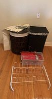 Photo showing two woven laundry hampers, one cylindrical dark brown with cream liner, one rectangular dark brown with black and white liner with lid open. Plastic box containing red velvet hangers with clips and white metal two-tier shoe holder rack placed in front.