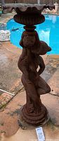 Full view of mother and child statue with planter top near poolside, showing overall shape and rusted metal stand.