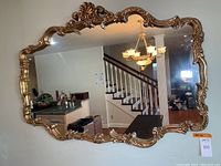 Front view of large rectangular wall mirror with ornate gold carved wood frame hanging on a white wall, reflecting a chandelier and stairway.