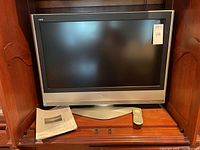 Front view of Panasonic 26-inch LCD TV with remote and manual placed below the TV screen on a wooden surface, set inside a cabinet.