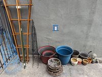 Pots in various sizes and colors and metal saucers stacked on the ground, with plant positioners and trellises leaning against the wall.