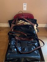 Photo shows a stack of eight women's purses in various colors and styles, mostly medium to large size, arranged on a flat surface for display.