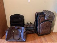 Four travel bags arranged on hardwood floor: leopard print large suitcase, leopard print smaller matching bag, black rolling suitcase, and brown overnight bag.