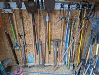 Wide view of the yard tools arranged in a shed, showing shovels, pickaxes, hoes, and rakes with various handle colors and signs of use.