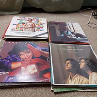 Four stacks of vinyl records on a carpet displaying multiple album covers including British Royal Family memorabilia from the XI Commonwealth Games and albums by Carole King, Janet Jackson, and Wham!.