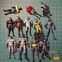 Photo front view of all 13 Batman action figures laid out on green cutting mat for scale and display showing details and colors.