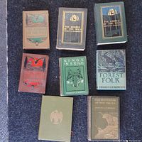 Top-down photo of 8 antique books by Charles G.D. Roberts arranged on dark fabric surface showing various cover colors and designs.