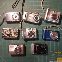 Front view showing lens and body of eight digital cameras on green grid mat